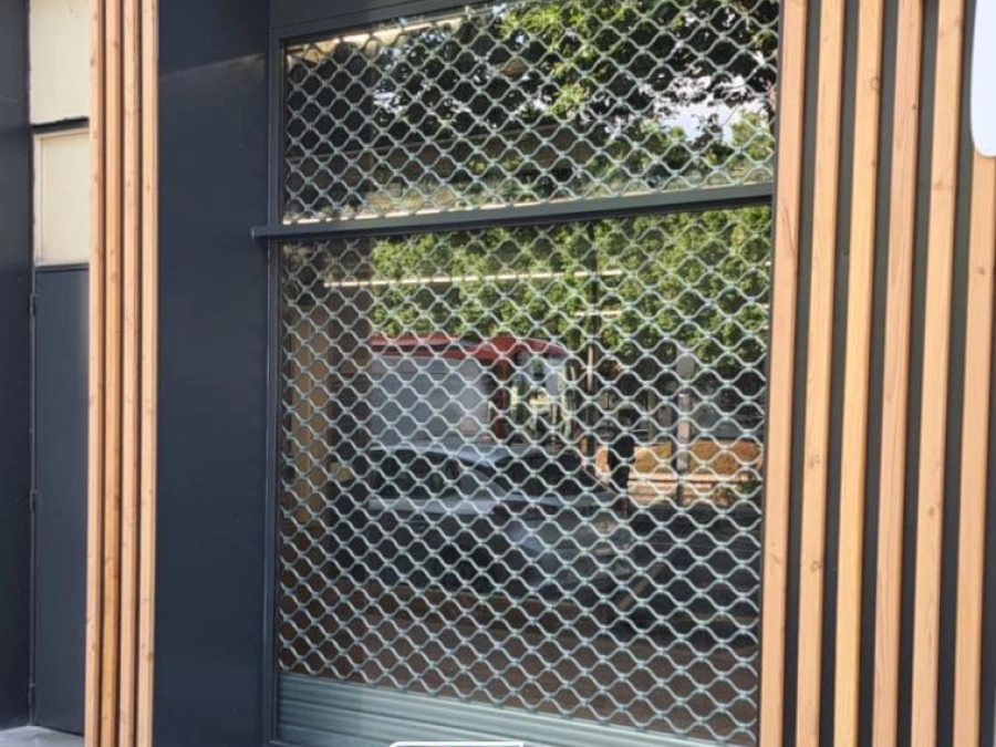 Grille de sécurité métallique à mailles sur devanture commerciale anthracite et bois, pose AFC à Caen.