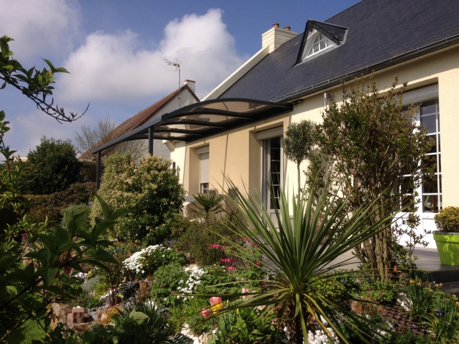 Abri de terrasse aluminium noir à toit cintré translucide, jardin fleuri d'une maison en Normandie