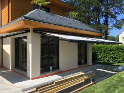 Stores extérieurs anthracite déployés sur baies vitrées d'une extension, jardin et pins en Normandie